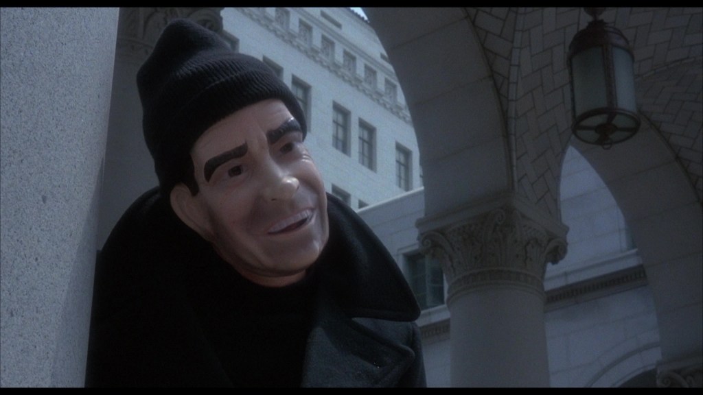 A figure wearing a black beanie and jacket, with a mask resembling a man's face, stands near an architectural column, framed by a historic building in the background.