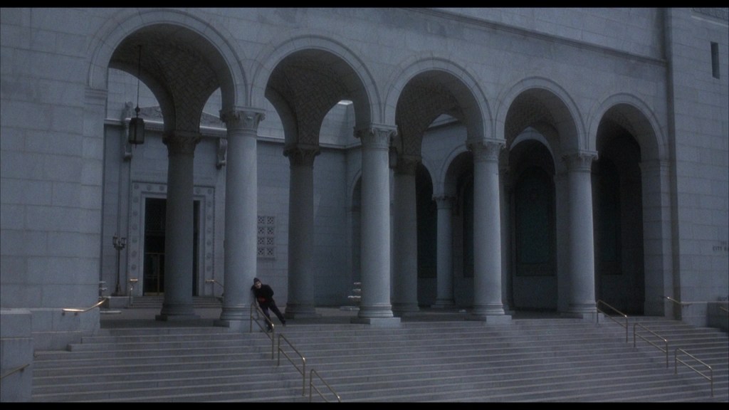 A person leaning against a column on the steps of a large, classical building with archways and ornate ceilings.
