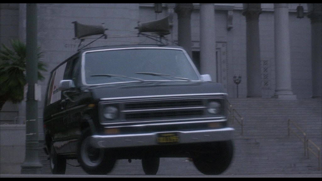A black van with loudspeakers on top jumps in front of a building's steps.