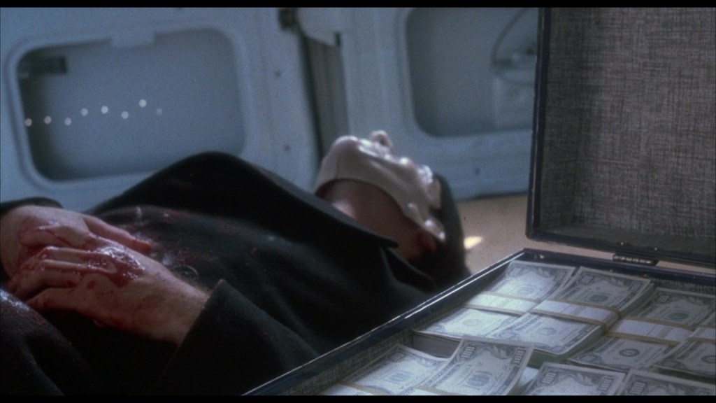 A man lying on the ground with a bloodied hand and a briefcase filled with cash open beside him.