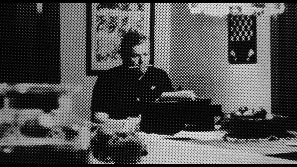 A person sitting at a table with a typewriter, appearing focused while writing. The setting includes some decor and a plate on the table.