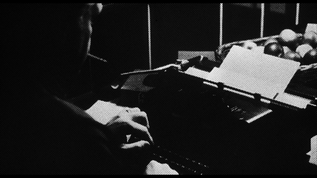 A person typing on a typewriter in a dimly lit room, with a sheet of paper and various objects in the background.