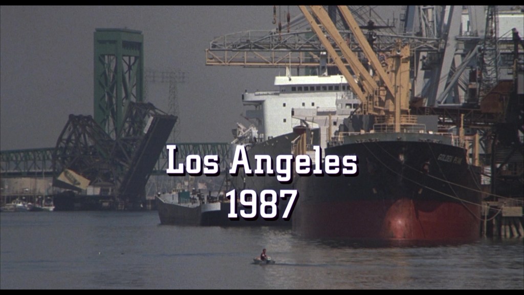 A waterfront view of Los Angeles in 1987, featuring industrial ships and cranes, with a small boat on the water.
