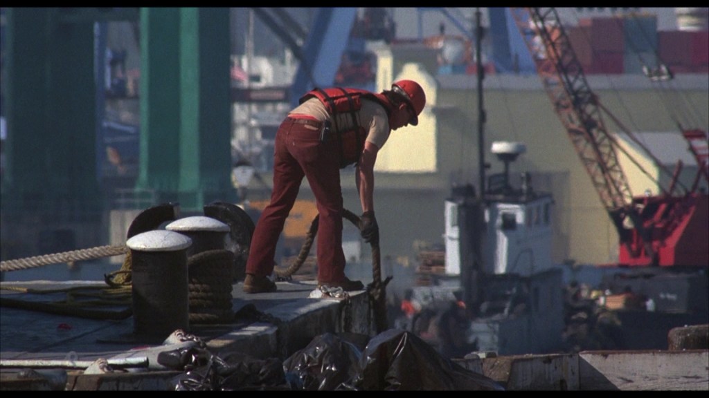 A worker wearing a hard hat and life vest is bending over the edge of a dock, handling equipment near boats and cranes in a busy port area.