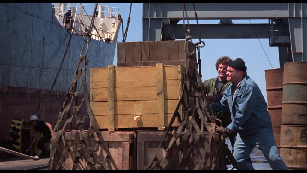 Workers loading wooden crates onto a ship at a dock, with barrels in the background.