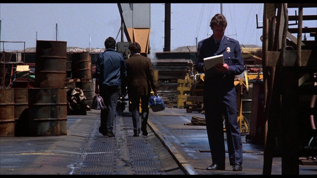 A police officer in a uniform holding paperwork stands on a dock, while two men walk away from him, passing by barrels and industrial equipment in the background.