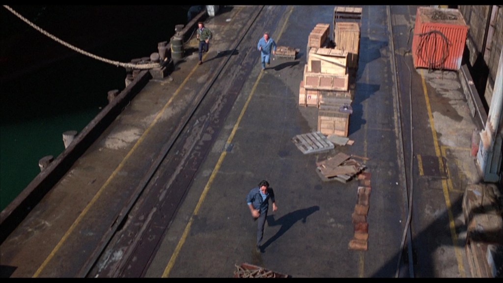 Three men running along a dock covered with shipping crates and debris, with a green body of water visible in the background.