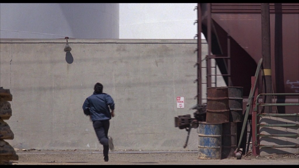 A person running away from a large wall in an industrial area, with barrels and a 'No Parking' sign visible nearby.