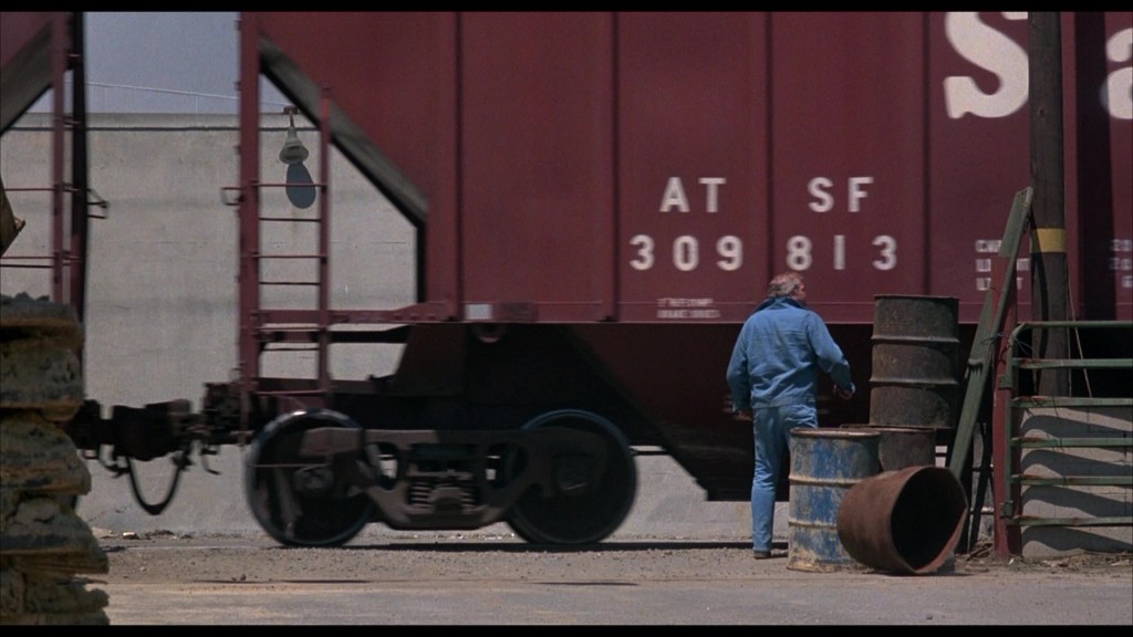 A man in a blue shirt and jeans walks near industrial barrels and a moving freight train, set against a backdrop of a gray wall.