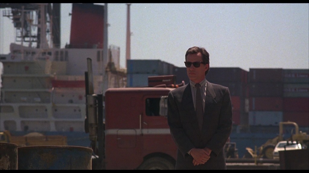 A man in a suit and sunglasses stands confidently in front of a shipping yard, with cargo containers and a large ship in the background.