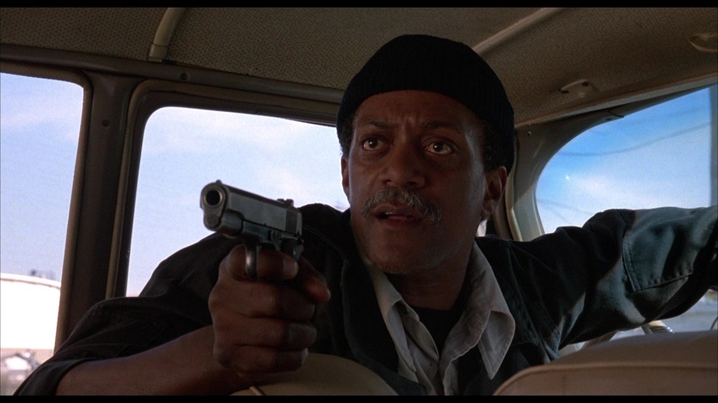 A man wearing a beanie holds a handgun while seated inside a vehicle, displaying concern and intensity in his expression.