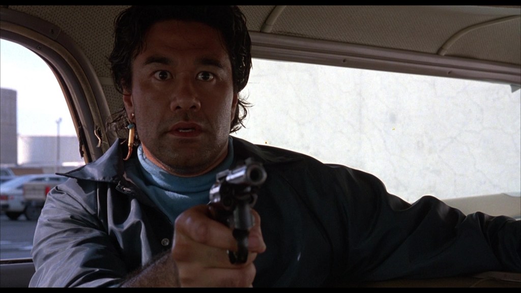 A man holding a handgun while sitting in a car, looking seriously at something outside the window.