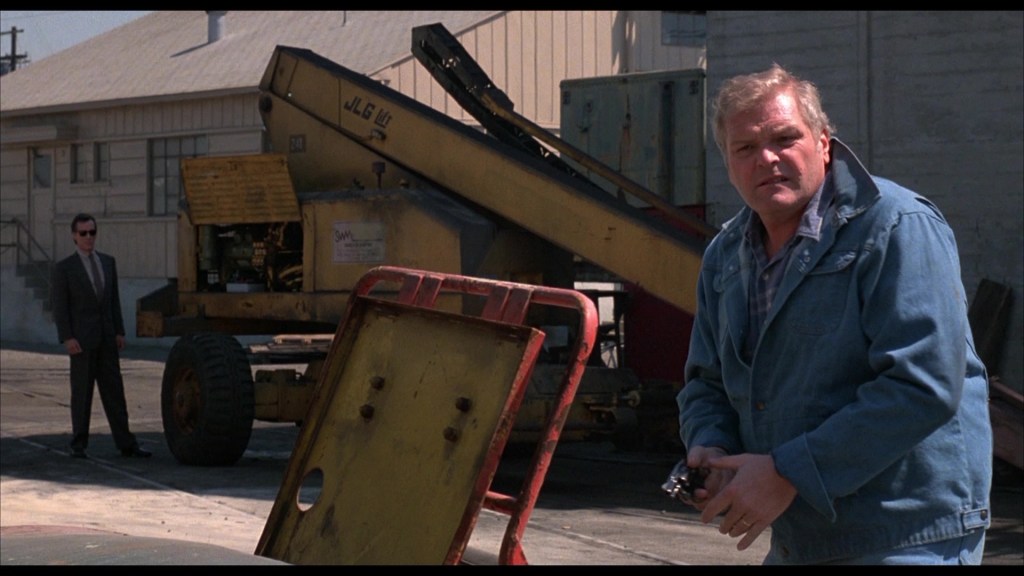 A man in a blue jacket stands holding a camera in front of a yellow industrial machine, while another man in a suit watches from a distance.