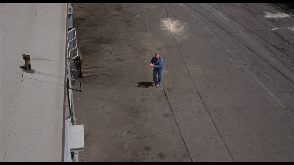 Aerial view of a man in blue clothing standing on a deserted street, with buildings and a visible open window alongside.
