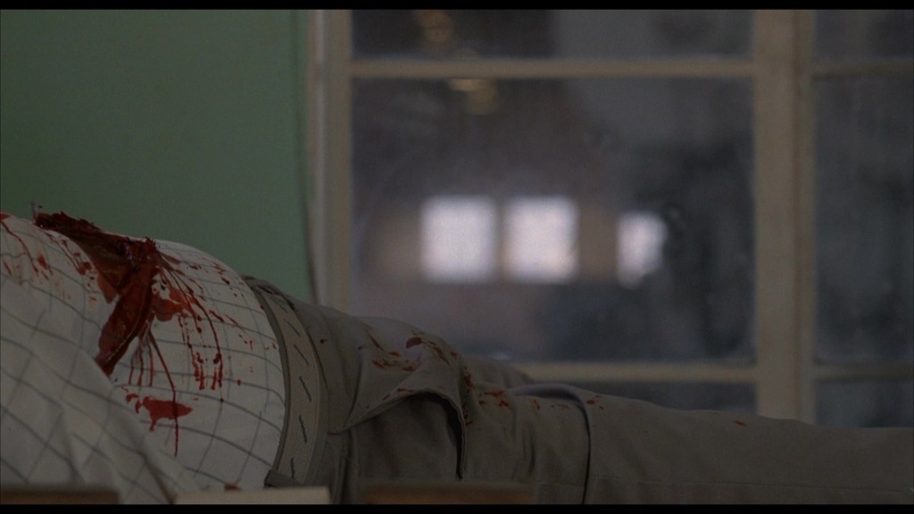 A close-up of a blood-stained shirt on a body lying on a surface, with a blurred background showing a window.