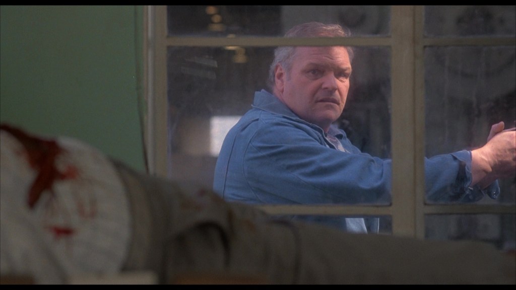 A man in a blue shirt stands behind a window, looking intently, while a figure in the foreground appears to be injured with blood on their clothing.