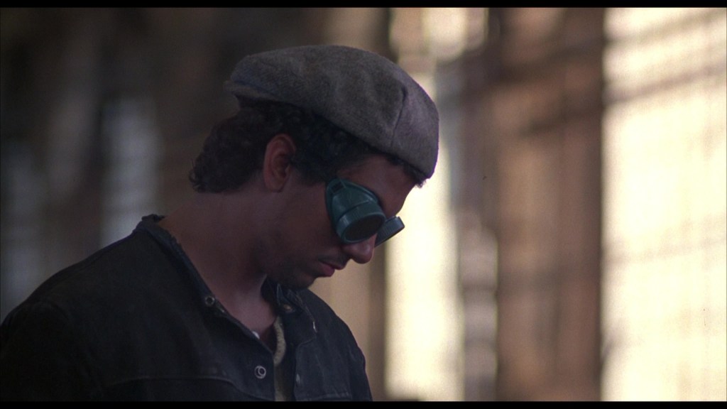 A person wearing a gray beret and teal sunglasses, looking down thoughtfully in a dimly lit space.