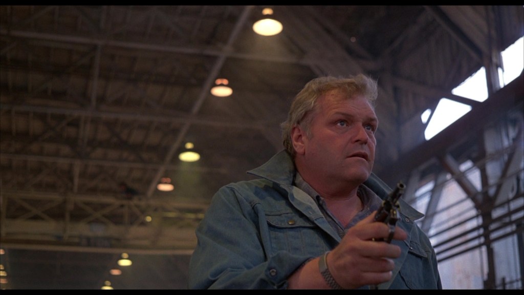 A man in a blue jacket holding a firearm, standing in an industrial location with high ceilings and soft lighting.