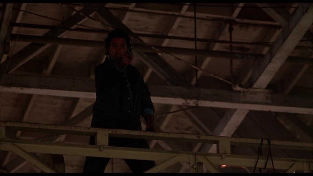 A man standing on a catwalk in a dimly lit warehouse, pointing a gun.