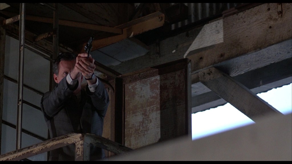 A man in a suit aims a gun from an elevated position in an industrial setting.