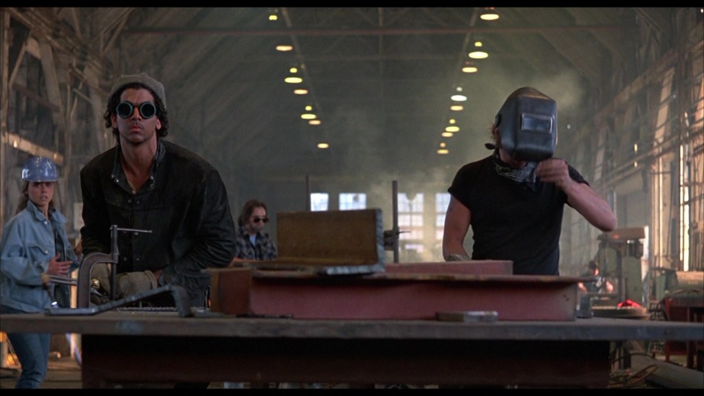 A group of workers in a dimly lit industrial warehouse, one wearing goggles and another with a welding helmet, focused on their tasks while smoke and light filters through the space.