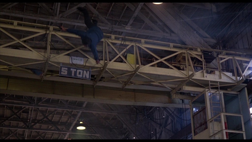 A person is seen falling from a high industrial beam inside a warehouse, with a sign reading '5 TON' visible above.
