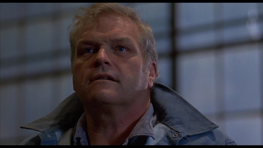 Close-up of a middle-aged man with short hair, wearing a blue jacket, looking up with a concerned expression in an industrial setting.