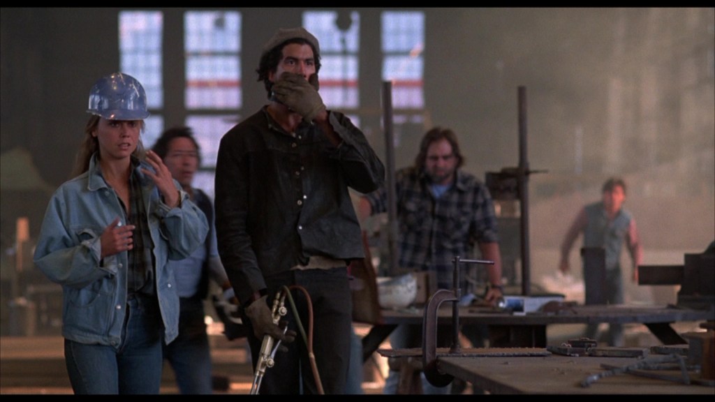 A group of people in a workshop setting, with one woman wearing a hard hat and looking concerned, while a man covers his mouth in shock. The environment appears industrial with tools and equipment visible.