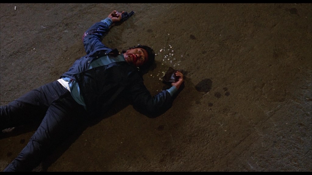 A man lying on the ground with a gun in one hand and blood on his face, surrounded by scattered debris.