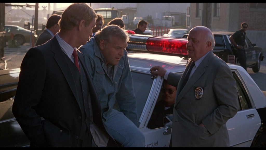 Three men discussing near a police car with flashing lights in an urban setting, with additional law enforcement in the background.