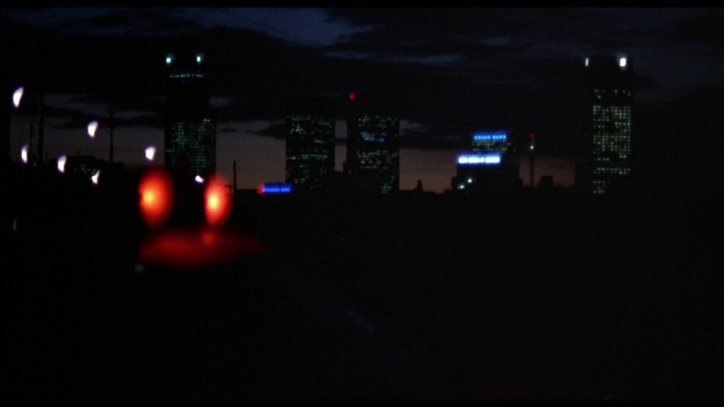 A nighttime cityscape featuring dark silhouettes of skyscrapers with a few illuminated windows and signage, juxtaposed against a twilight sky.