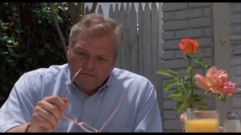 A middle-aged man in a light blue shirt looks thoughtfully at a pair of glasses he is holding, sitting at a table with a glass of orange juice and a vase of flowers in the foreground.