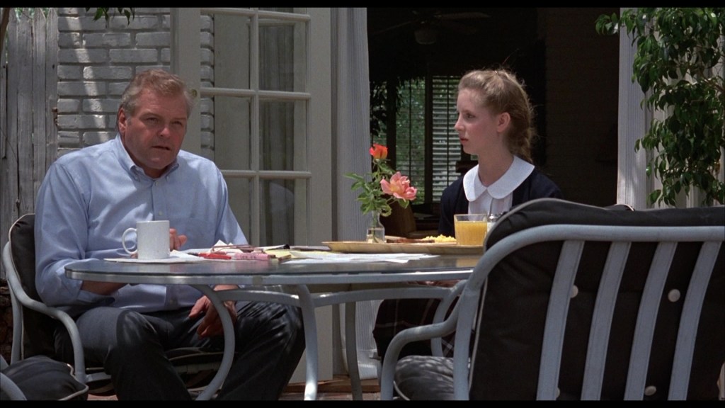 A man and a young woman are seated at a table outside, having breakfast. The table is set with plates of food and drinks, with a flower in a vase. The man looks concerned while the woman appears pensive.