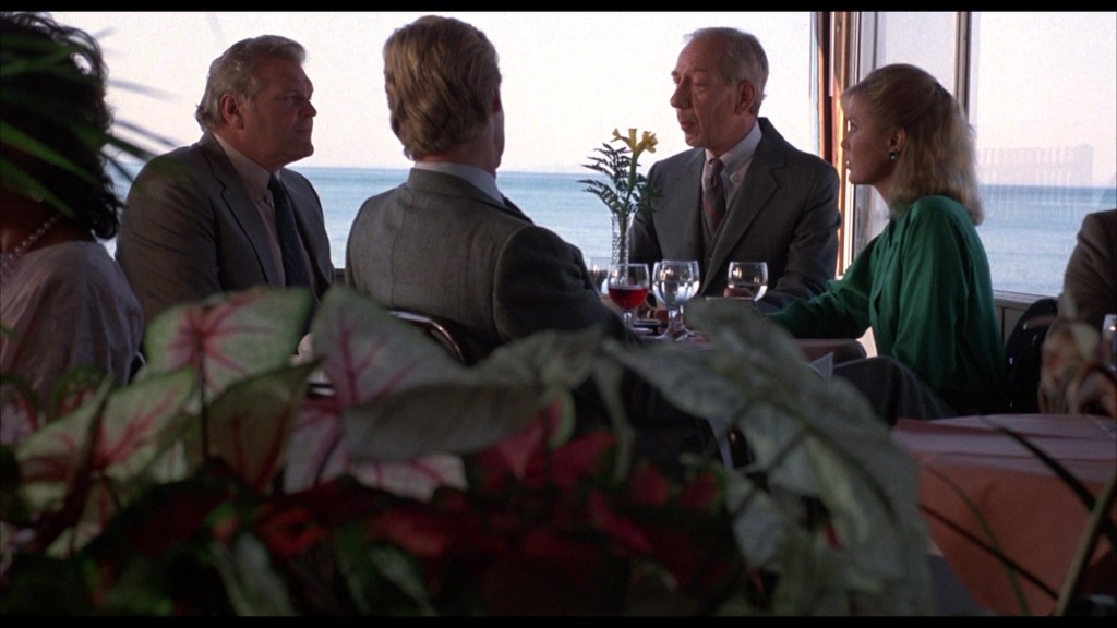 A group of four people seated at a restaurant table with a view of the ocean. The scene includes two men in suits, a woman in a green dress, and another man in a lighter suit. They appear to be engaged in conversation, with drinks on the table and tropical plants in the foreground.