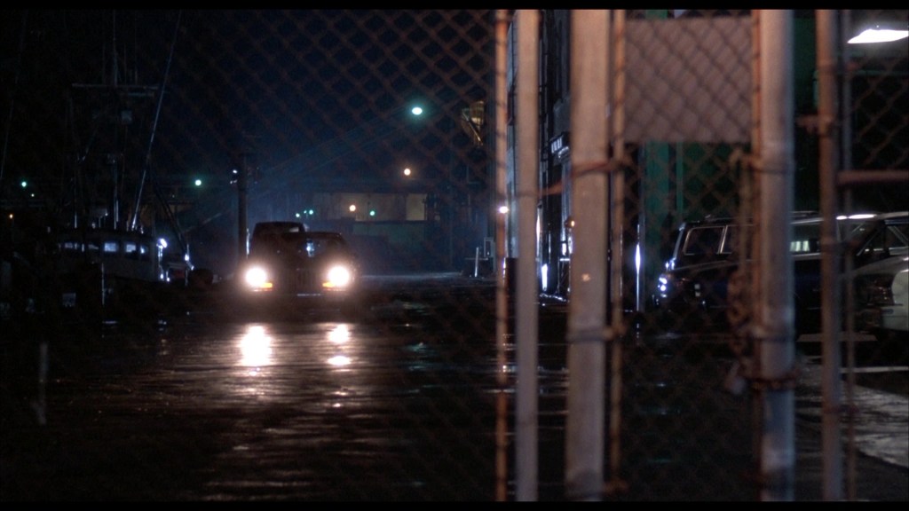 A nighttime scene showing a dimly lit street with a car approaching through a gated area, revealing reflections on the wet pavement and industrial buildings in the background.