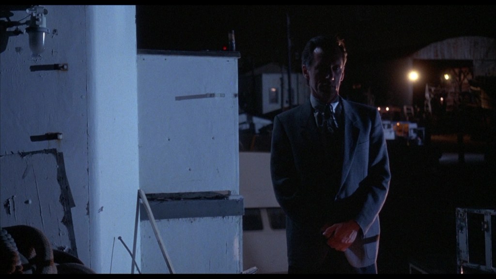 A man in a suit stands in a dimly lit outdoor setting at night, with a partially visible boat and buildings in the background.