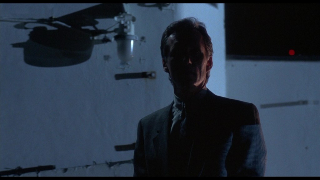 A shadowy figure stands in front of a white wall, with dim lighting creating a mysterious atmosphere.