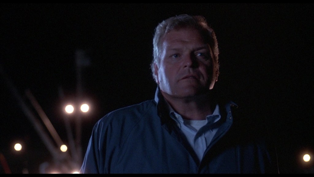 A man in a dark blue jacket stands under dim lights at night, appearing solemn and pensive.