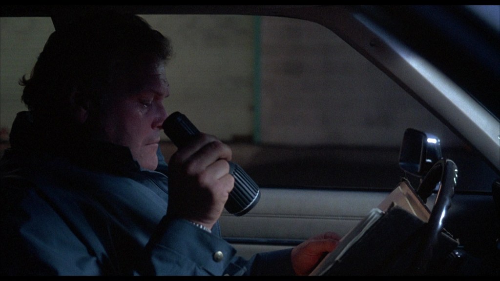 A man sitting in a car at night, holding a flashlight and looking at a clipboard.