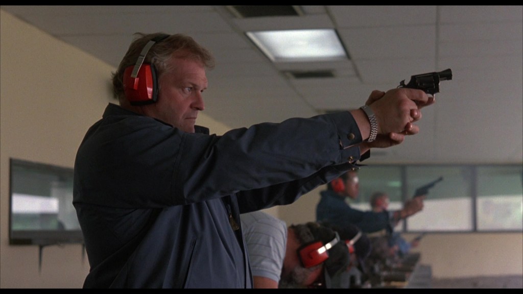 A man at a shooting range wearing ear protection aims a handgun at a target, with other shooters visible in the background.