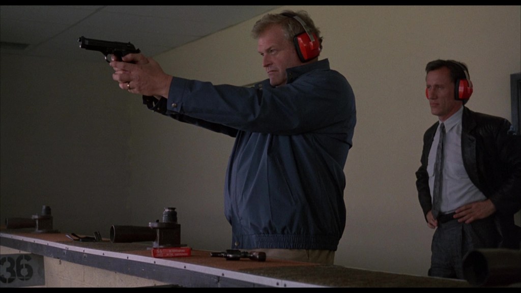 A man wearing ear protection aims a handgun at a shooting range, while another man in a suit observes him from behind.