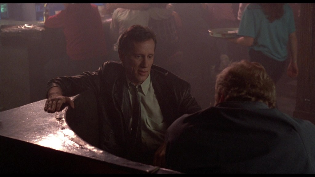 A man in a leather jacket sitting at a bar, engaged in conversation with another man whose back is facing the camera, in a dimly lit bar setting.