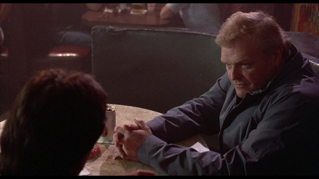A close-up of a man sitting at a table in a dimly lit bar, looking intently at another person off-camera. He has a serious expression and short, light-colored hair.