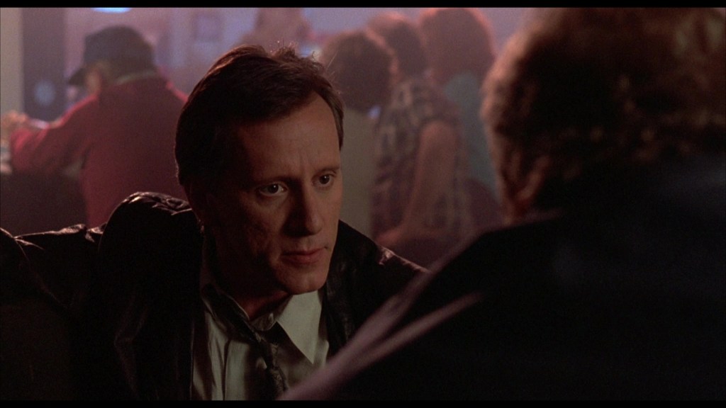 A close-up of a man with a serious expression, wearing a shirt and tie, engaged in conversation in a dimly lit bar setting.
