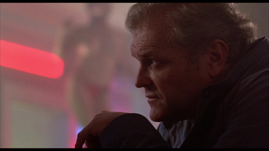 Close-up profile of a man with light gray hair and a serious expression, set against a softly lit background with red and pastel colors.