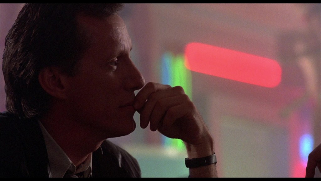 A close-up profile of a man with dark hair and a suit, thoughtfully resting his chin on his hand, with colorful neon lights in the background.