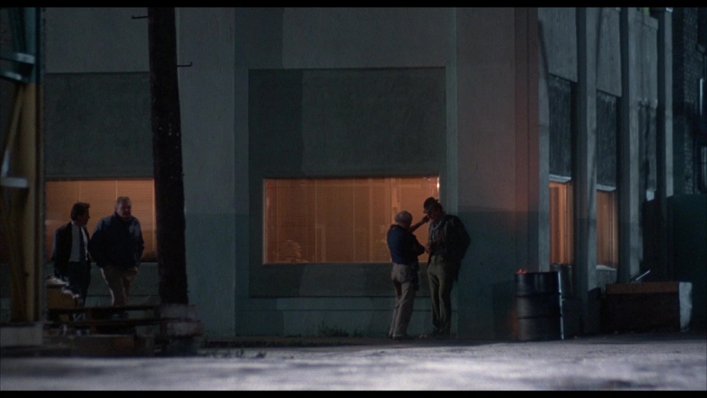 Two men are conversing in the foreground near a building at night, while another pair of individuals walks in the background. The building features large windows with warm lights glowing from inside.