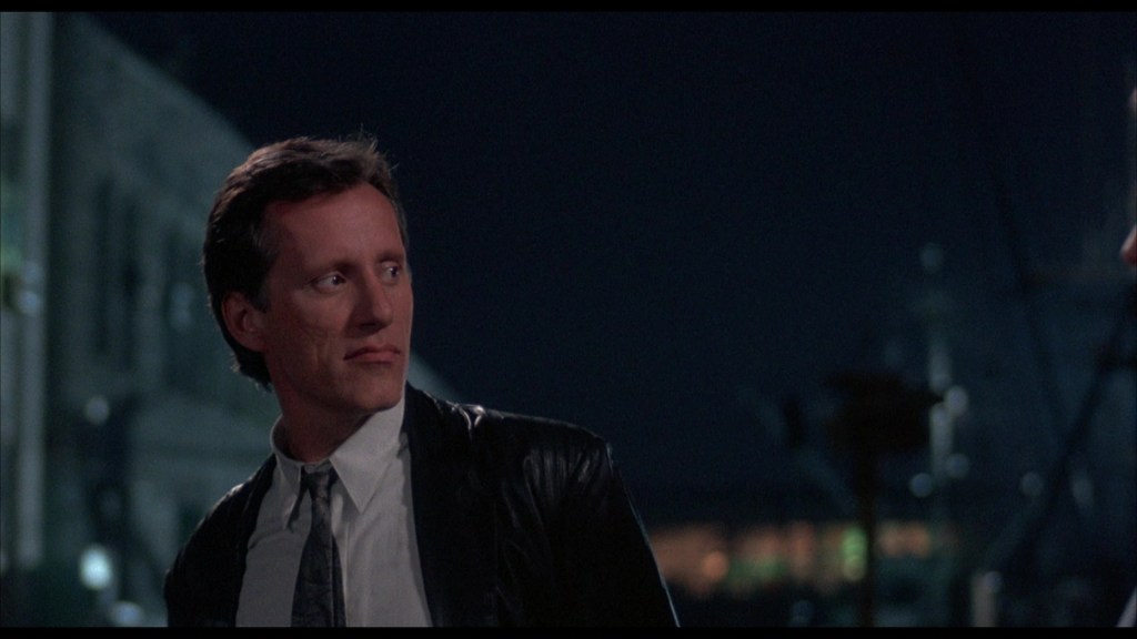 A man in a leather jacket and tie stands in a nighttime urban setting, looking thoughtfully to his right.