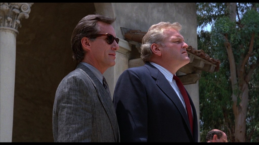 Two men in suits standing outside, one wearing sunglasses, looking up with serious expressions, against a backdrop of trees and a building structure.