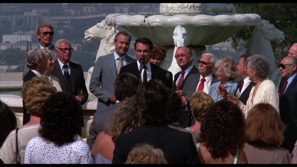 A group of people gathered around a man speaking at an outdoor event, with a fountain in the background.